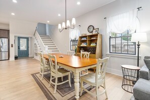 Un salón amplio con una mesa de comedor de madera, sillas blancas, una escalera, una estantería y una ventana con cortinas.