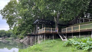 Ein Steg aus Holz am Seeufer mit überdachter Sitzgruppe und einer Treppe zu einer erhöhten Plattform.