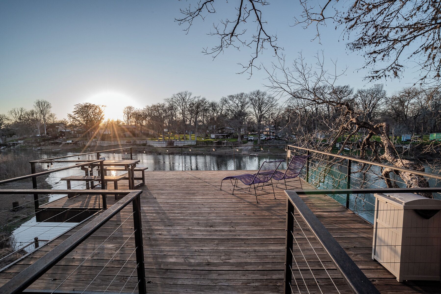 Una terraza de madera con mesas de picnic y una silla de descanso con vista a un cuerpo de agua, con un atardecer de fondo.