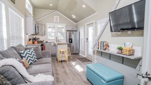 A compact kitchen with a refrigerator, sink, and a small island. There is a sofa and a blue ottoman in the living area. - River Escape / Fishing Dock / Kayak / FastWiFi (Seguin)