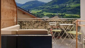 A balcony with a wicker sofa, a small table, and a chair, overlooking a scenic valley with houses and a church.