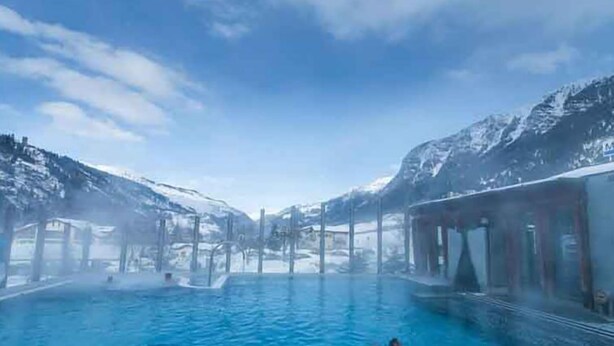 A pool with people swimming, surrounded by snow-capped mountains and a clear sky.