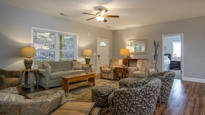 A living room with a sofa, armchairs, ottomans, a coffee table, lamps, a ceiling fan, and a door leading to another room. - BK's Hideaway, with fire-pit and bike storage (Pisgah Forest)