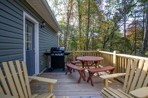 Une terrasse avec des chaises et des tables en bois, un barbecue et une maison dont le revêtement extérieur est de couleur sombre.