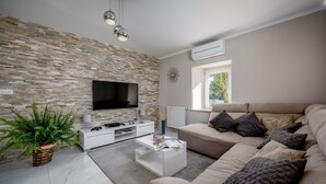 Uma sala de estar moderna com parede de pedra, televisão de tela plana, móvel branco para entretenimento, sofá bege com almofadas estampadas, planta em vaso e janela com vista para o verde.