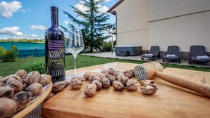 Uma garrafa de vinho, um copo e nozes sobre uma mesa de madeira ao ar livre, com um edifício e cadeiras ao fundo.