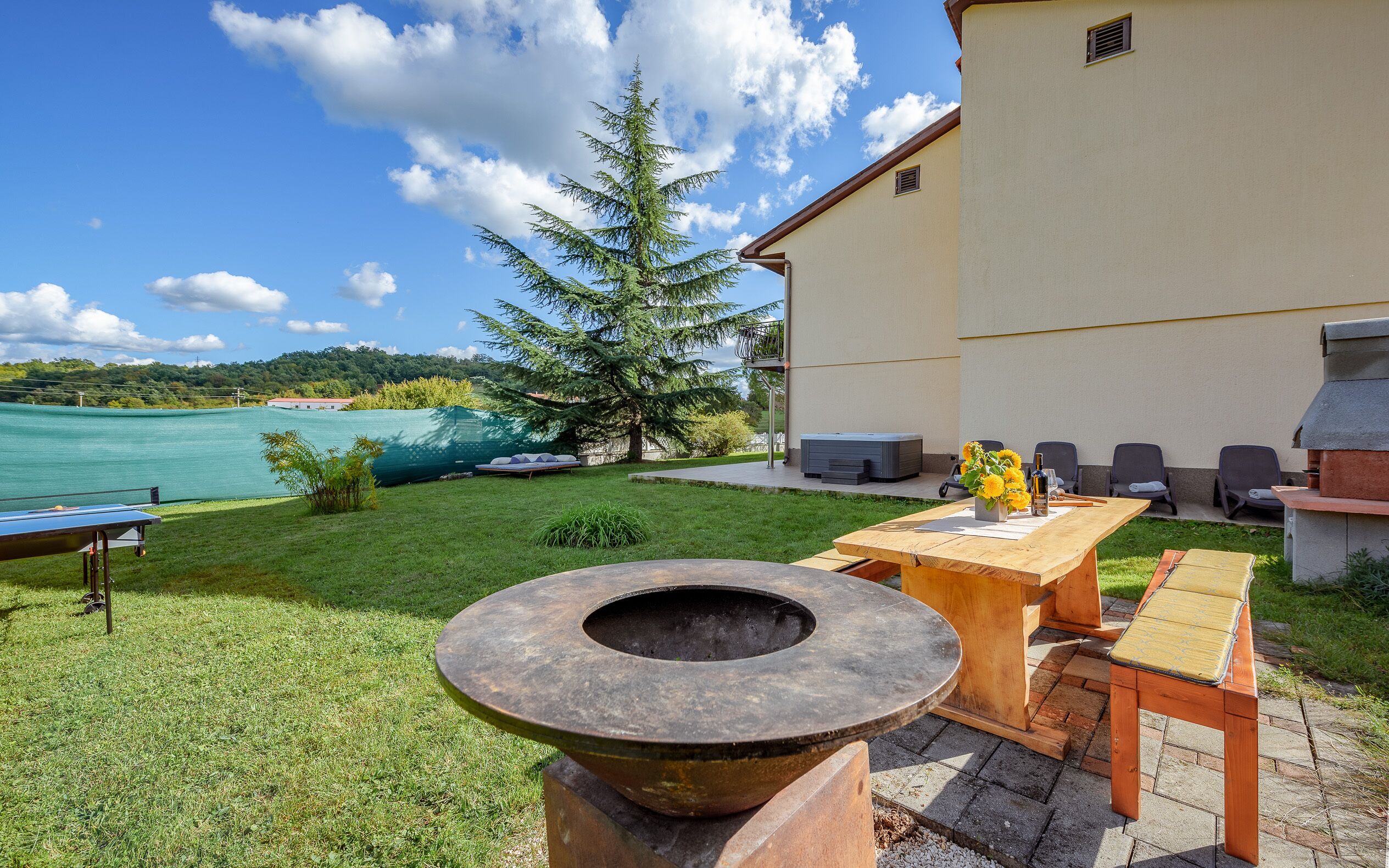 Eine Terrasse im Freien mit einem Steinkamin, einem Holztisch und Stühlen, mit Blick auf einen See und einen großen Baum.