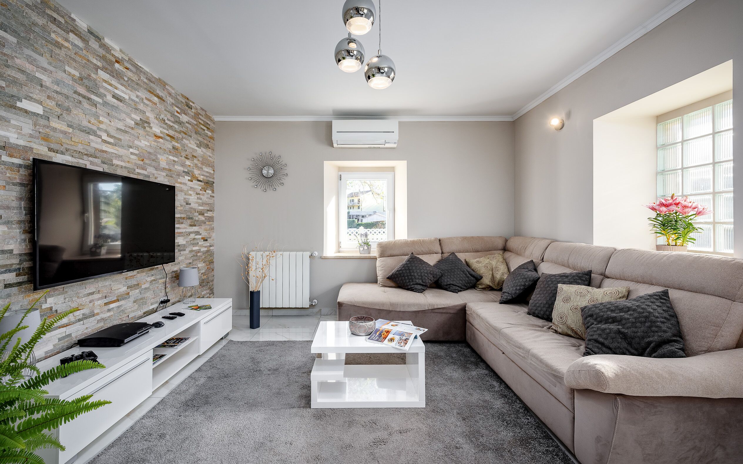 A modern living room with a flat-screen TV, a sectional sofa, a white coffee table, and a stone wall.