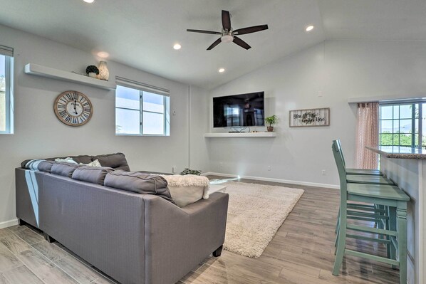 A living room with a grey sofa, a ceiling fan, a flat-screen TV, a clock, and a window.