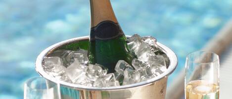 A bucket with a bottle and ice, two glasses of champagne, and a cork on a table by a pool.