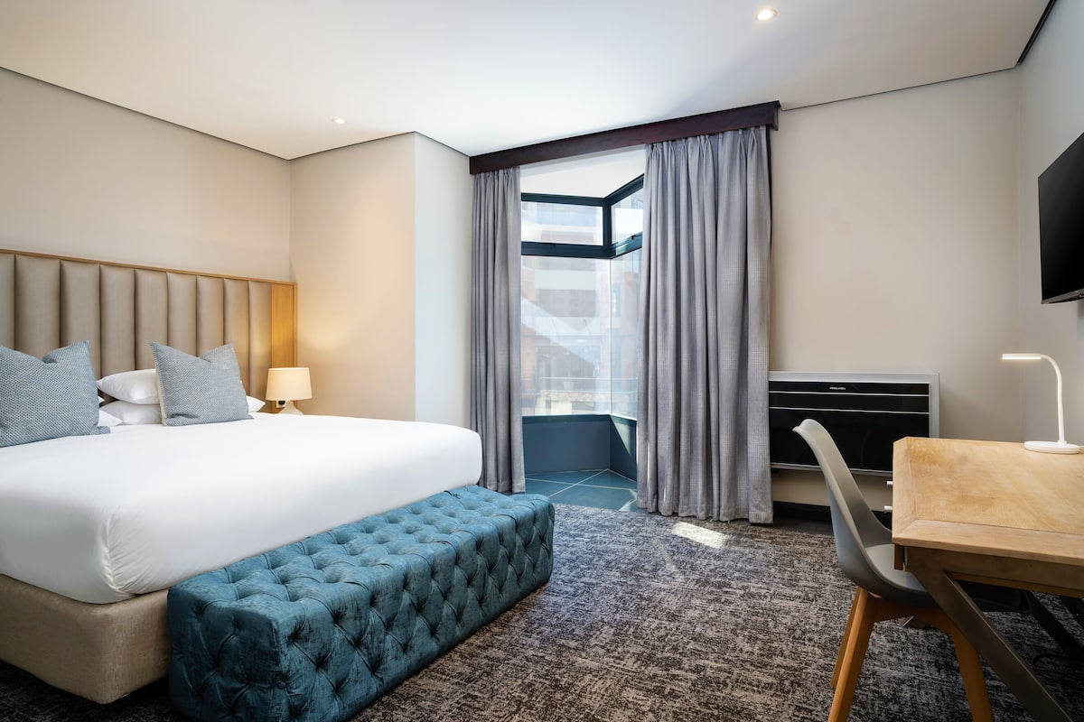 A hotel room with a large bed, a bench, a desk, a flat-screen TV, and a view of the city through a window.
