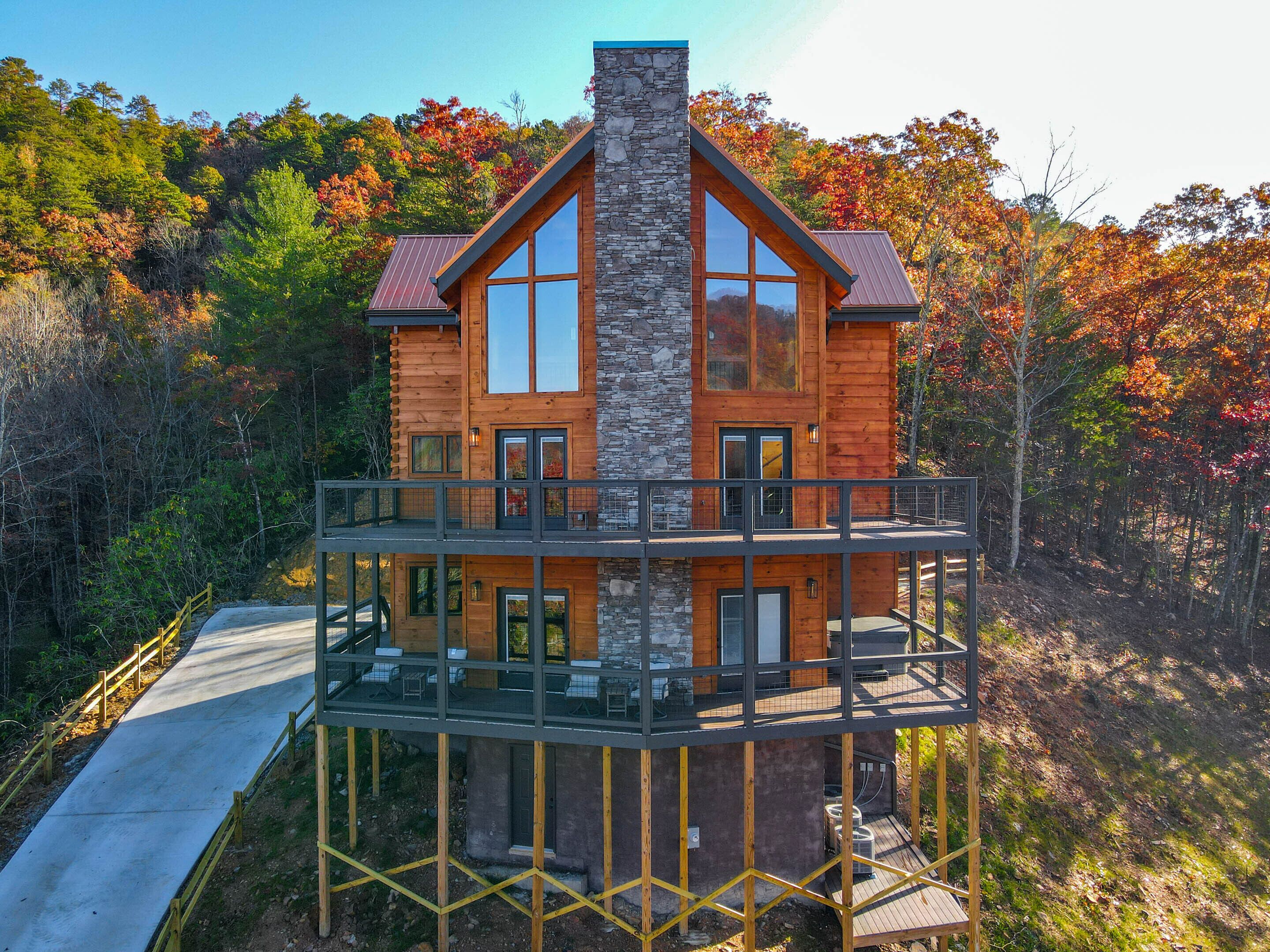 A two-story log cabin with a stone chimney, a balcony with outdoor seating, and a scenic forest backdrop.