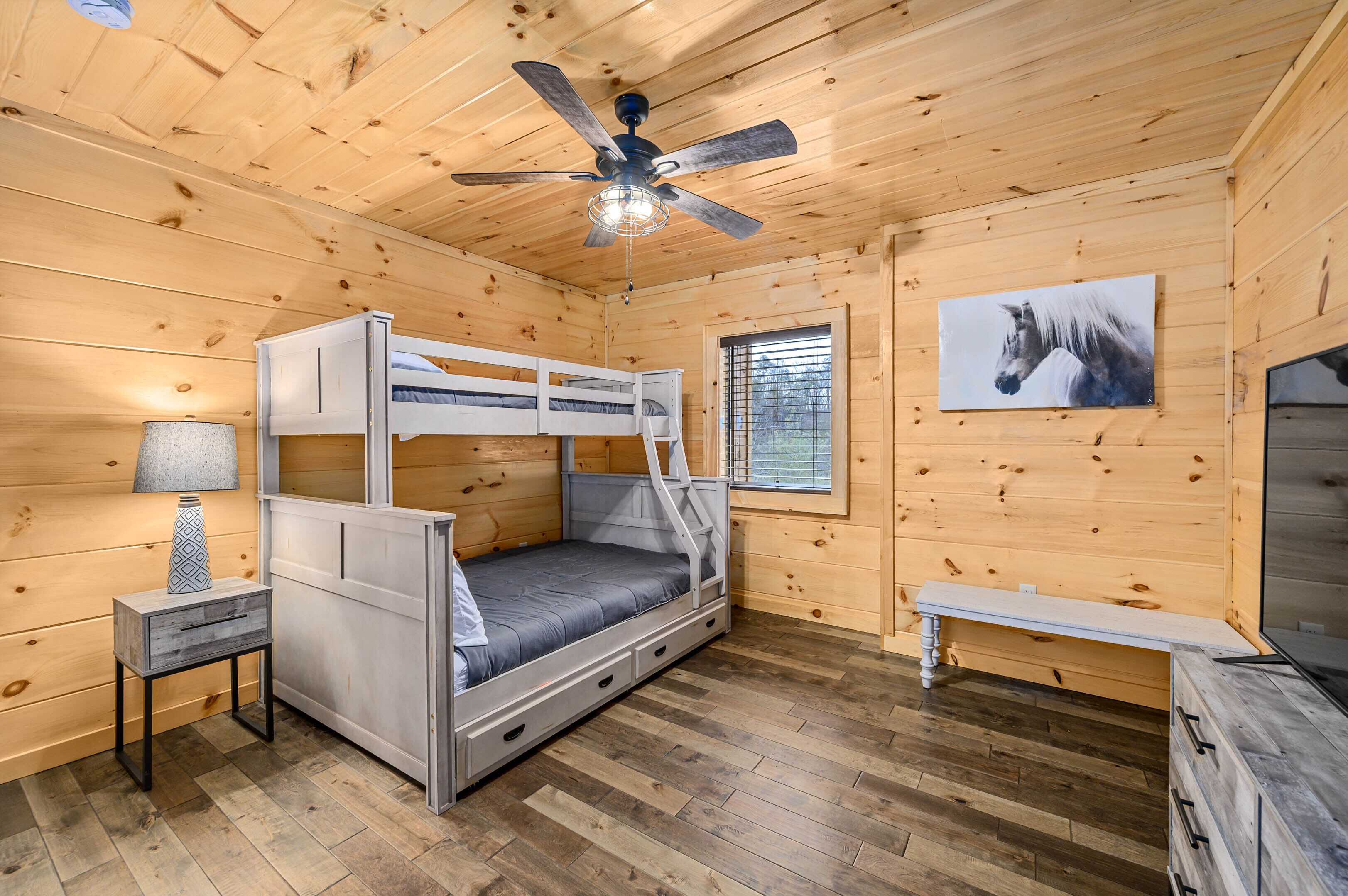 Una habitación de cabaña de madera con literas, ventilador de techo, ventana con persianas, lámpara y un cuadro de un caballo en la pared.