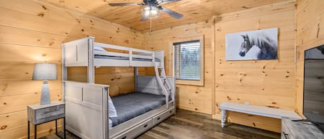 Una habitación de cabaña de madera con literas, ventilador de techo, ventana con persianas, lámpara y un cuadro de un caballo en la pared.