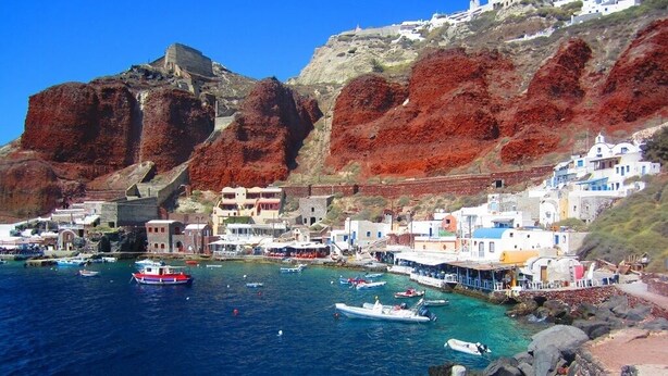 A coastal town with buildings on a hillside, boats in the water, and rocky shores. - Room-pleasure Room 2 By The Sea (Santorini)