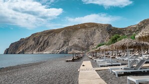 A beach with sun loungers and thatched umbrellas, a wooden walkway, and a rocky cliff in the background. - Room-pleasure Room 2 By The Sea (Santorini)