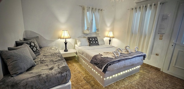 A bedroom with a bed, a sofa, two bedside tables with lamps, a window with curtains, and a door with a sign.