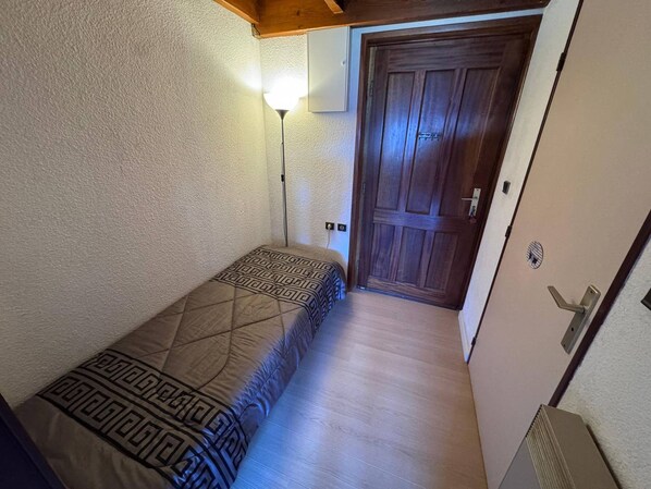 Una habitación pequeña y estrecha con una cama individual, una puerta de madera y una lámpara fijada a la pared.