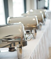 Una fila de estaciones de buffet con tapas y mangos metálicos, sobre un mantel blanco.