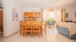 A dining area with a wooden table and chairs, a bookshelf with books and decorative items, and a view of the outdoors through a window. - Beach House Villa Isabel (Santanyí)
