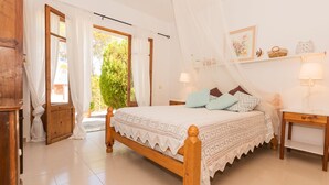 A bedroom with a wooden bed, white curtains, a bedside table with a lamp, and a window with a view of greenery. - Beach House Villa Isabel (Santanyí)