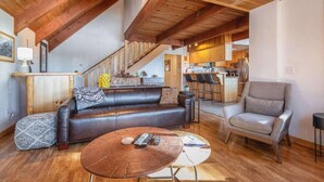 A cozy living room with a leather sofa, a round coffee table, and a grey armchair. A wooden staircase leads to a kitchen area with wooden cabinets and a bar counter. - Lakeview Serenity Cottage (Kings Beach)