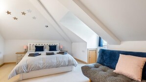 A bedroom with a sloped ceiling, a bed with striped bedding, a bedside table with a lamp, and a sofa with cushions. - Spacious Flat A Few Mins Walk to Carlyon Bay Beach (Carlyon Bay)