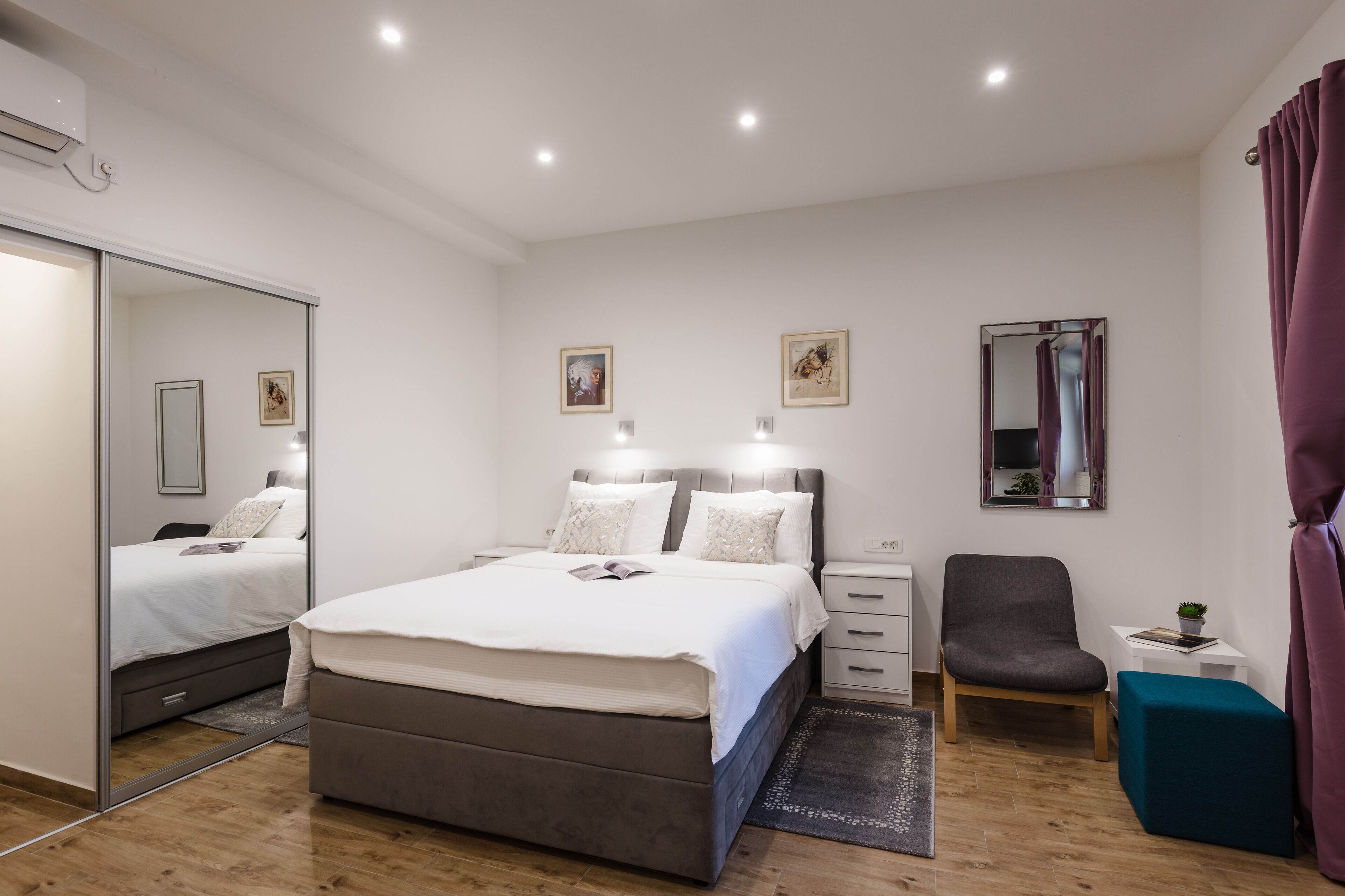 A modern bedroom with a large bed, bedside tables, a chair, a small table with a plant, and a mirror on the wall.