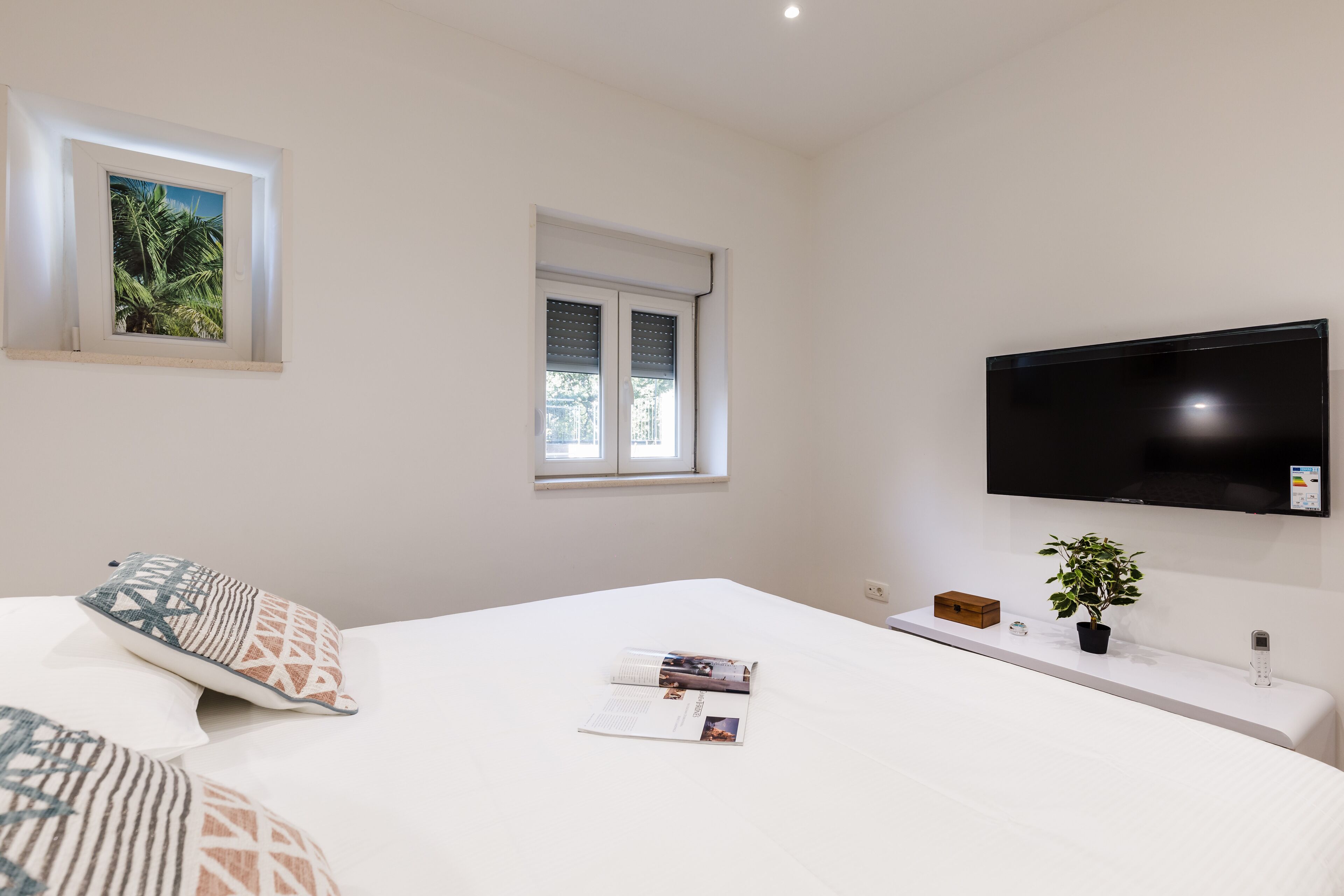 A neatly arranged bedroom with a bed, a TV mounted on the wall, a window with blinds, and a small plant on a bedside table.