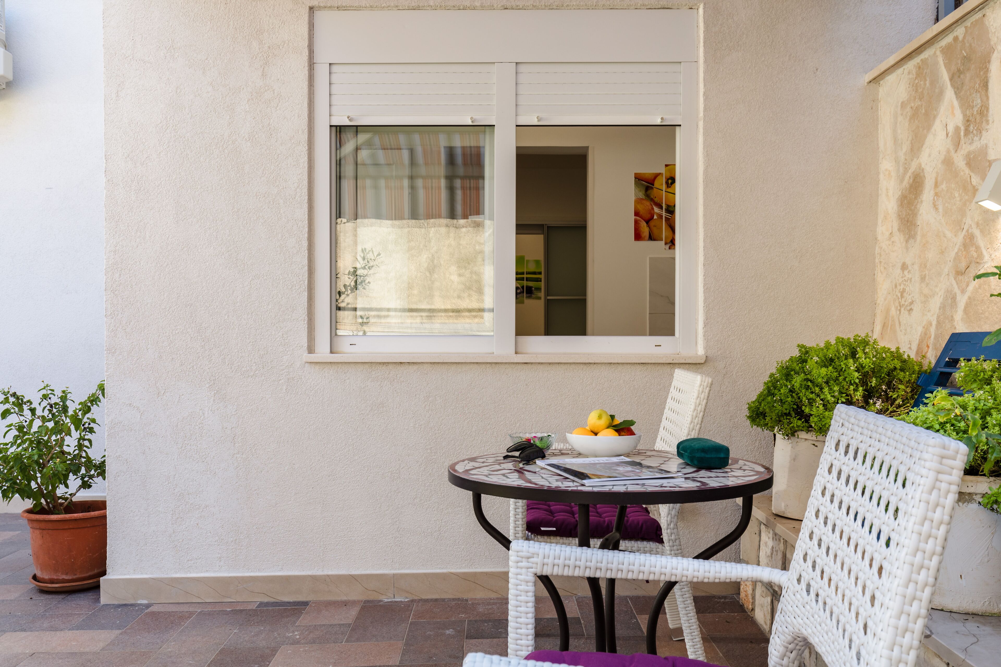 Un pequeño patio al aire libre con una mesa redonda, una silla de mimbre blanco y una planta en maceta.