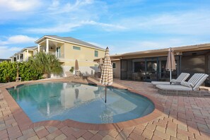 A swimming pool with a brick border, lounge chairs, and an umbrella.