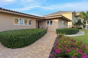A residential building with a paved walkway, a well-maintained hedge, and flowering plants.