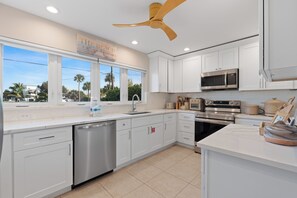 A modern kitchen with white cabinets, stainless steel appliances, and a large window overlooking palm trees.