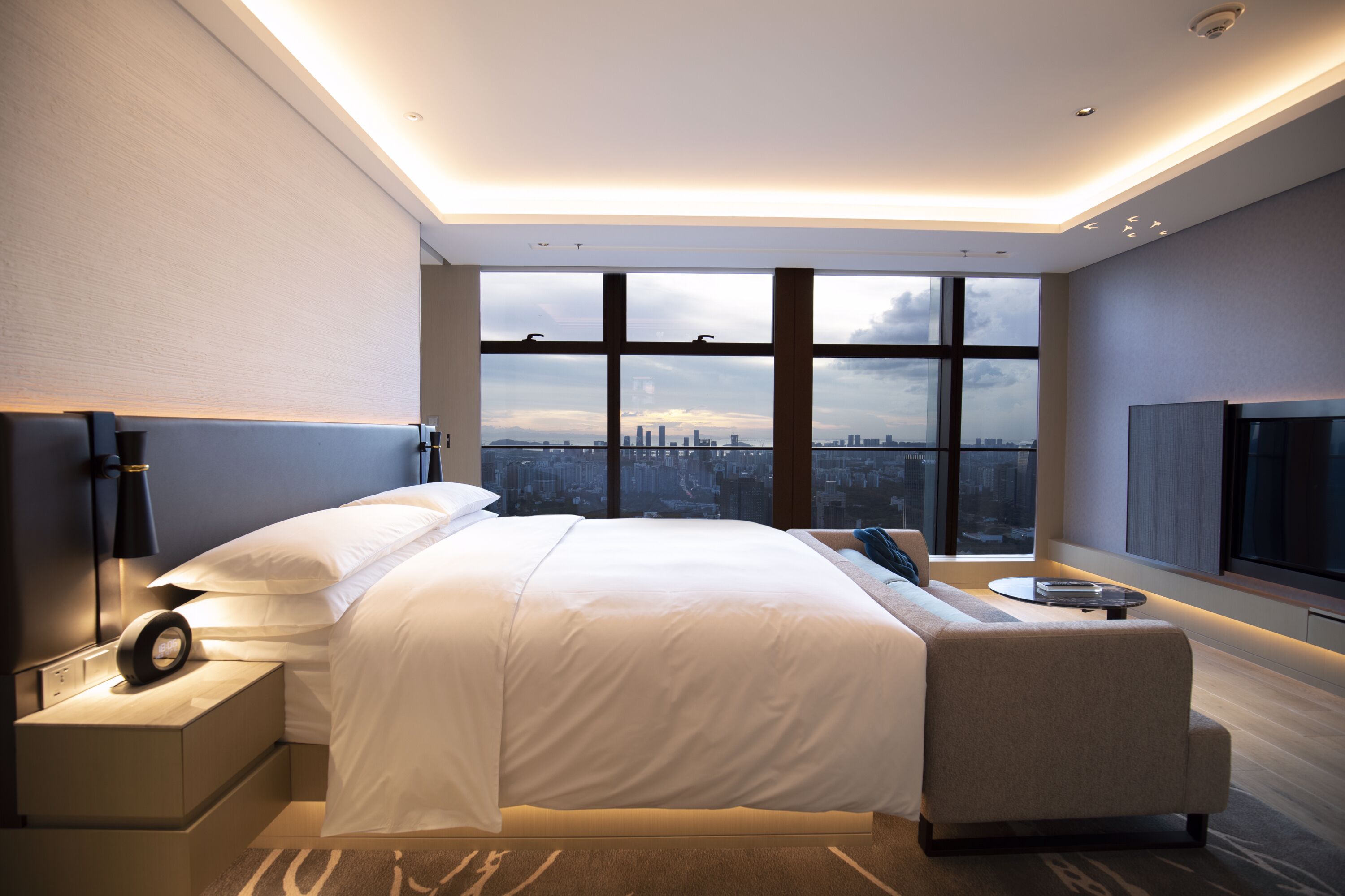 A modern hotel room with a large bed, a bedside table with a lamp, a view of the city through large windows, and a flat-screen TV mounted on the wall.
