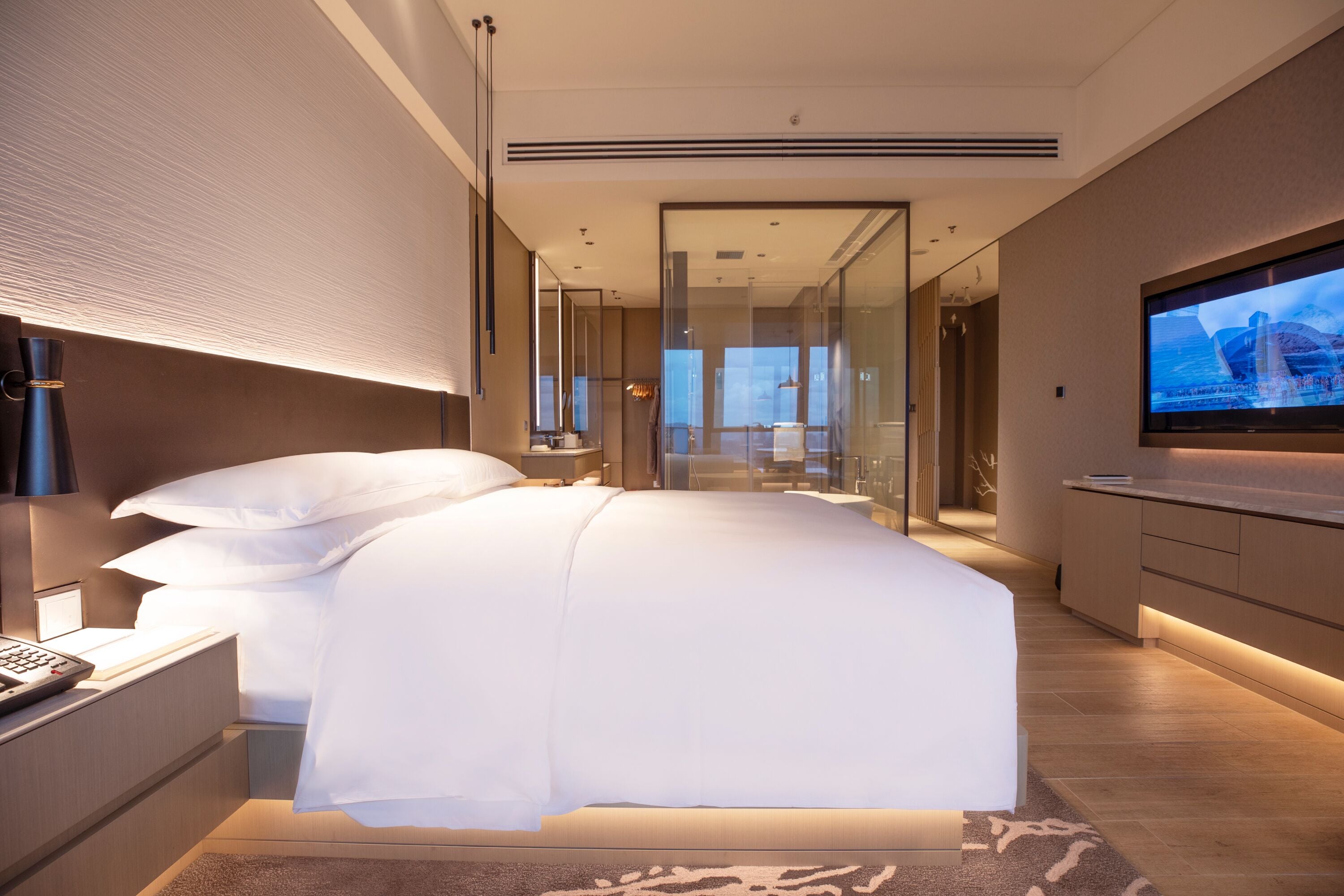 A modern hotel room with a large bed, a television, and a glass partition wall.