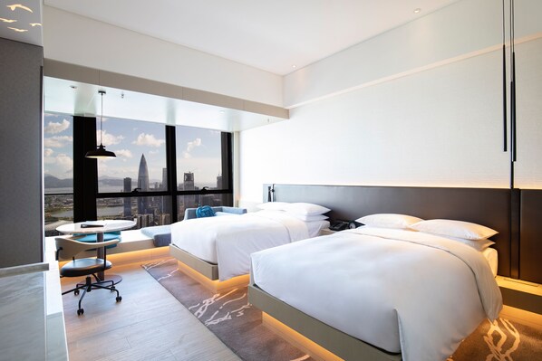Habitación de hotel moderna con dos camas, un escritorio y vistas a un paisaje urbano.
