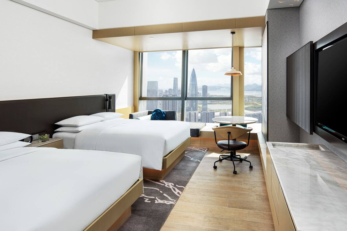 A hotel room with two beds, a desk, a chair, a TV, and a city view.