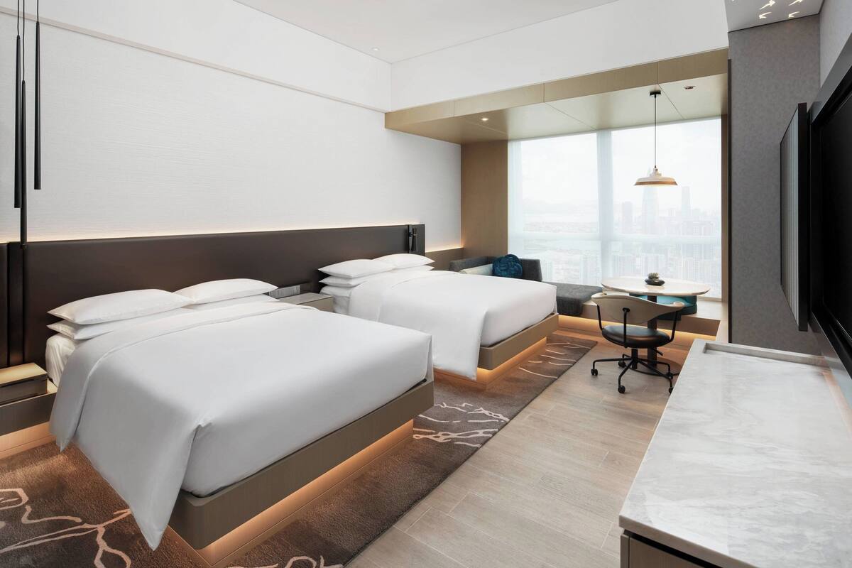 A hotel room with two beds, a desk, and a large window offering a city view.