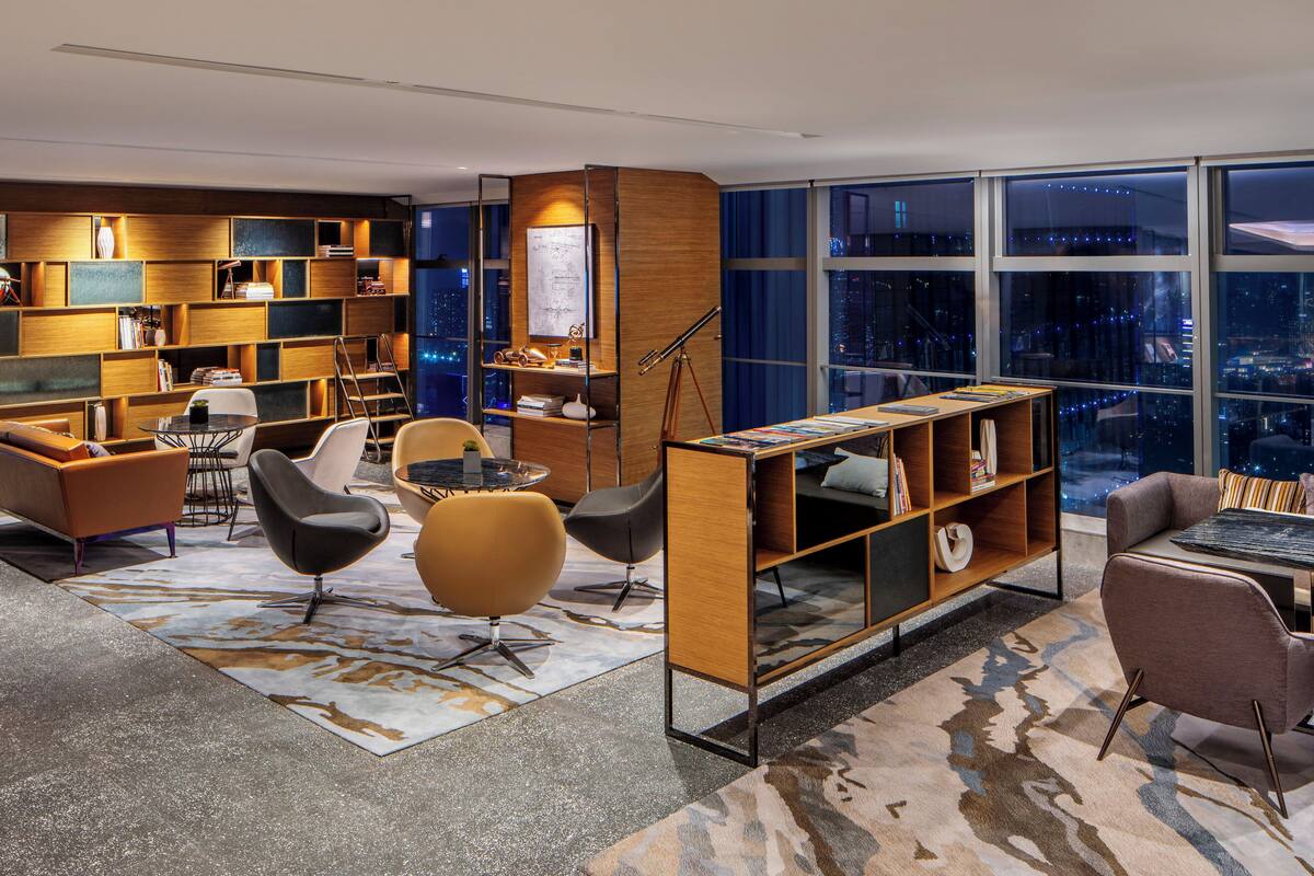 A modern lounge area with a city view, featuring a mix of seating options, a bookshelf, and a small table.