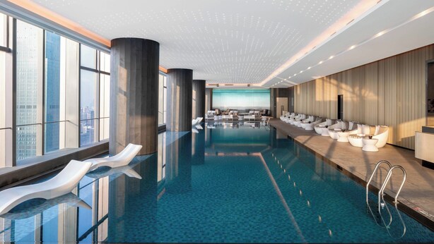 Una piscina interior moderna con agua azul cristalina, sillones de descanso y vistas a la ciudad a través de amplios ventanales.