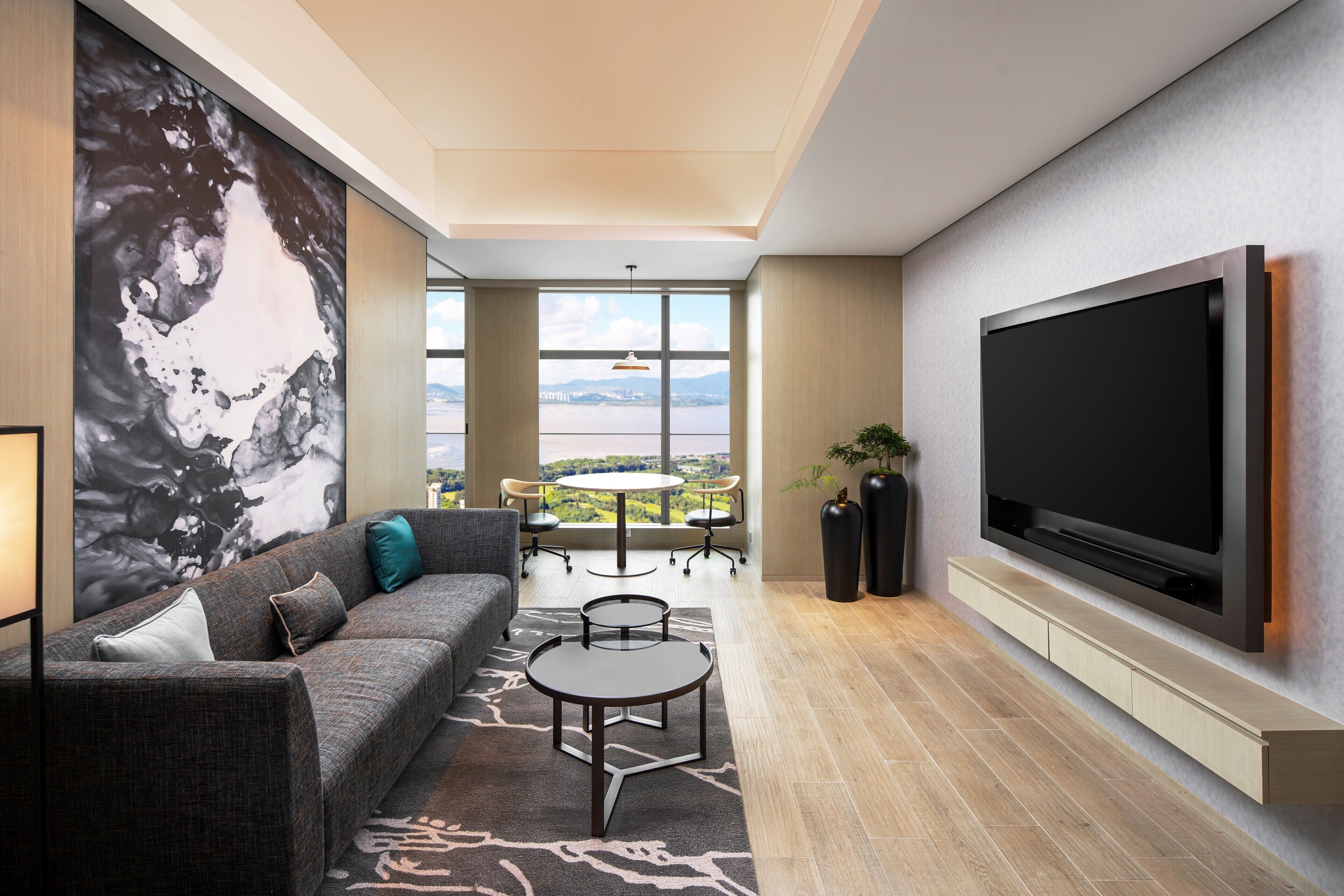 A modern living room with a large flat-screen TV, a gray sofa with teal cushions, a coffee table, and a view of a bridge and greenery through large windows.