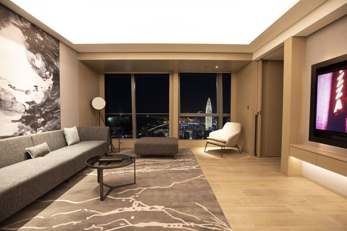A modern living room with a sofa, ottoman, and coffee table, large windows with a city view, and a flat-screen TV mounted on the wall.