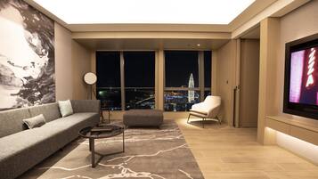 A modern living room with a sofa, ottoman, and coffee table, large windows with a city view, and a flat-screen TV mounted on the wall.
