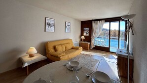 A cozy living room with a sofa, a dining table set for two, and a large window offering a view of the sea.