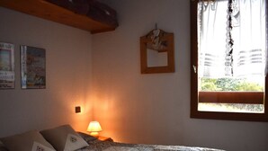 A bedroom with a bed, a nightstand with a lamp, a window with curtains, and wall decorations. - 2-Room Cabin Apartment with Parking in Font-Romeu (Font-Romeu-Odeillo-Via)