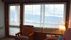 A room with a wooden chair and a sofa, a window with a view of mountains, and a lamp on a side table. - 2-Room Cabin Apartment with Parking in Font-Romeu (Font-Romeu-Odeillo-Via)