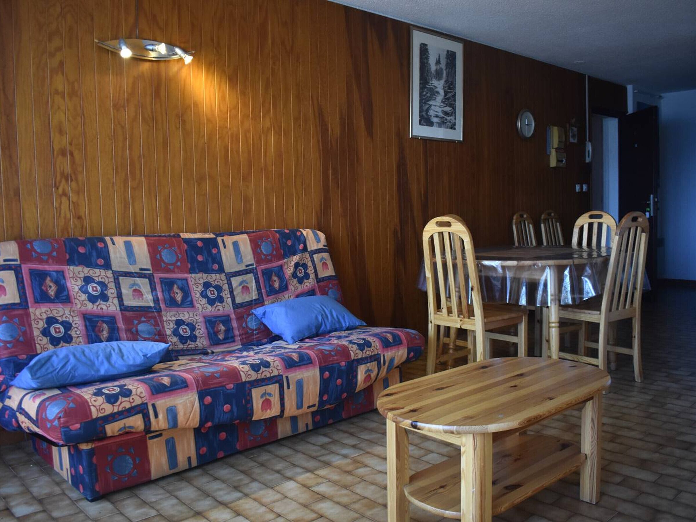 Een knus vertrek met een houten wand, een gestreept banksofa, een houten eettafel en stoelen.