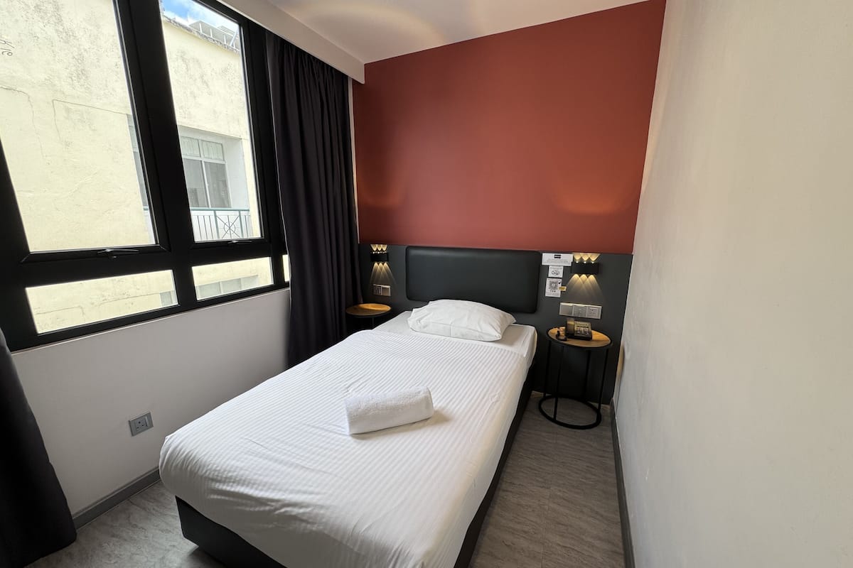 Habitación de hotel con cama, mesita de noche, lámpara de pared y ventana con cortinas.