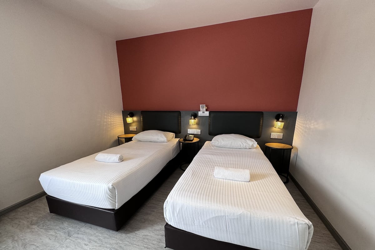 Habitación de hotel con dos camas individuales, cada una con una colcha blanca y una toalla enrollada. Se observan mesitas de noche con lámparas y un teléfono en la pared.