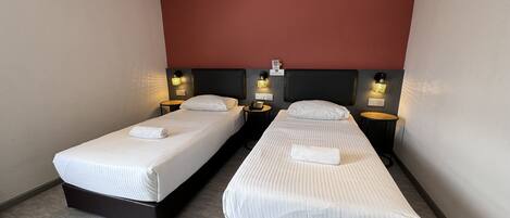 A hotel room with two single beds, each with a white bedspread and a rolled towel. There are bedside tables with lamps and a wall-mounted phone.